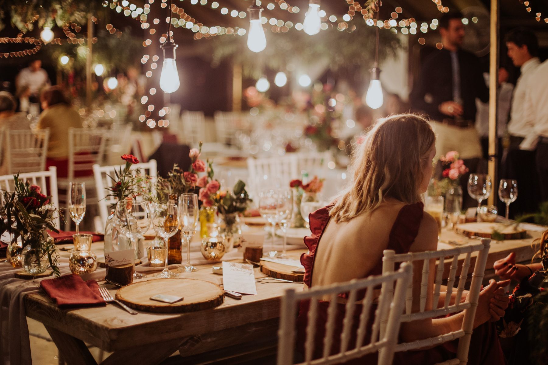 Atmospheric wedding portfolio image of young lady in open back maroon dress sitting at wedding reception with elegant wedding decor